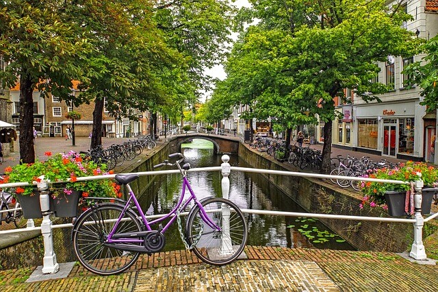 Eine kleine Brücke mit weißem Geländer über einem Kanal im niederländischen Delft, ein lilafarbenes Fahrrad lehnt an der Brücke, blühende Bäume säumen den Kanal und Blumenkästen mit roten Blumen hängen am Brückengeländer.