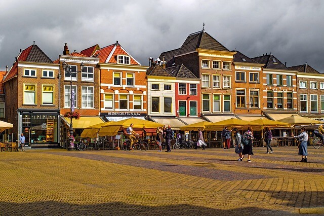 Ein Stadtplatz im niederländischen Delft mit einer Häuserfront, die von Sitzmöglichkeiten ansässiger Lokale mit gelben Sonnenschirmen gesäumt ist, die Sonne wirft die Häuserfront in glitzerndes Licht